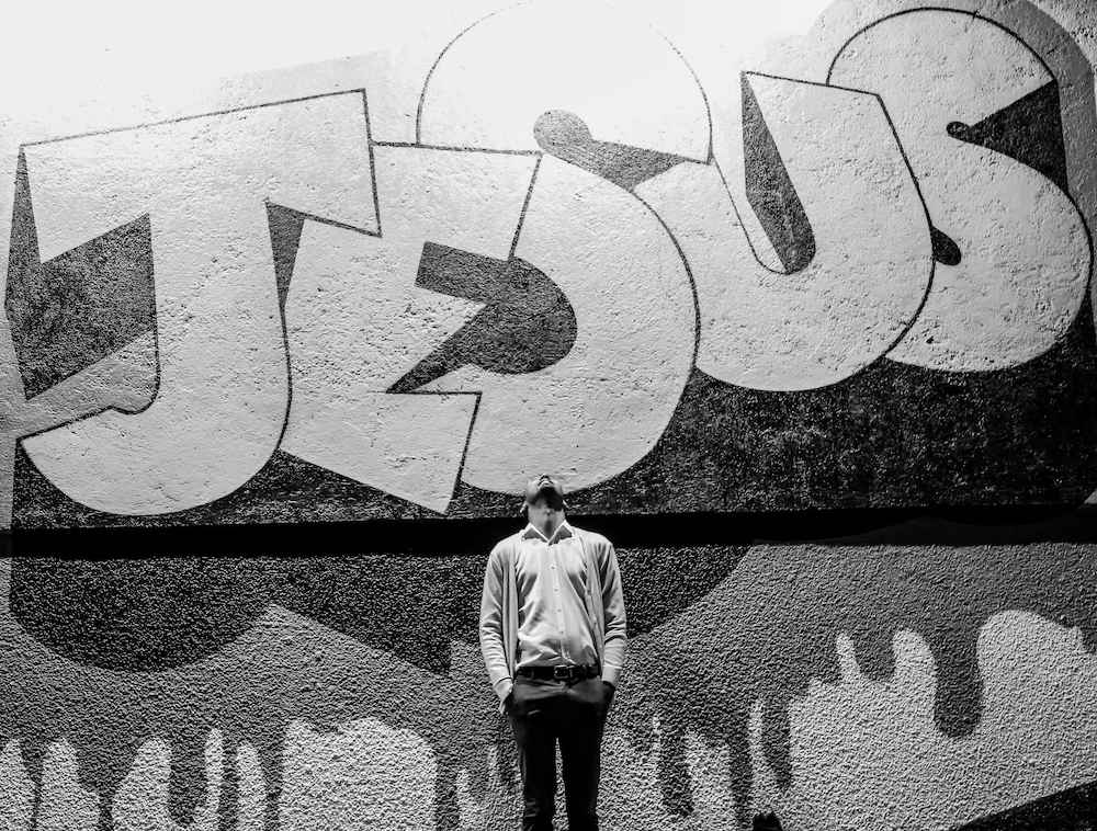 Guy standing underneath Jesus' name spray painted on wall.